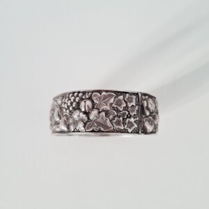 May include: A wide silver-toned metal cuff bracelet featuring a detailed embossed pattern of grape clusters and ivy leaves. The surface has a darkened, oxidized finish in the recessed areas to highlight the intricate botanical design.