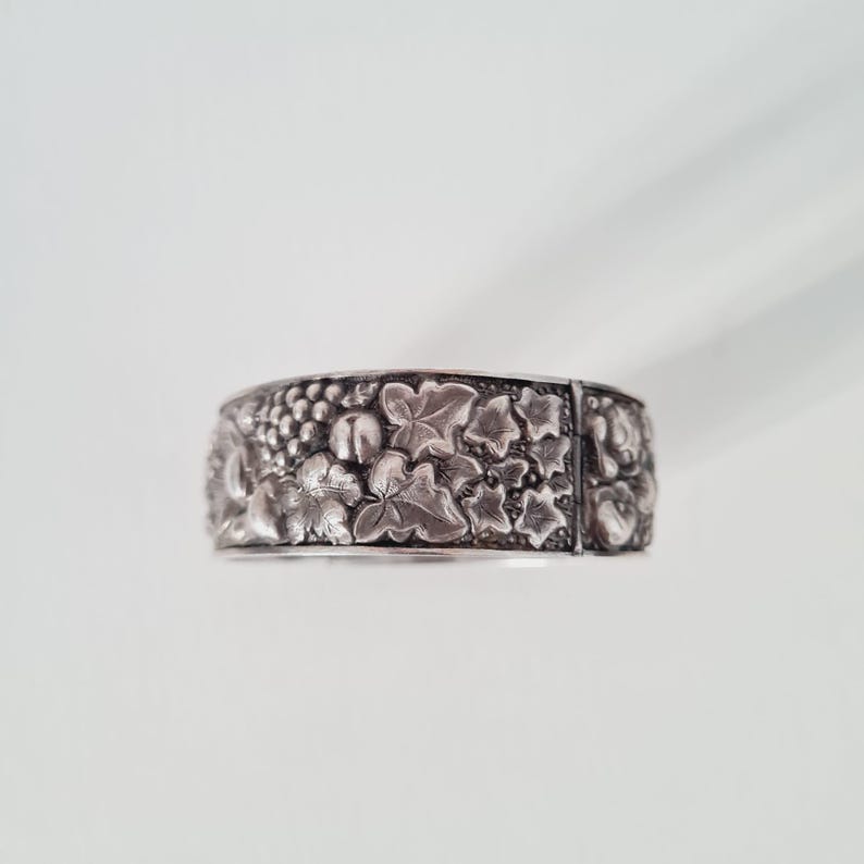 May include: A wide silver-toned metal cuff bracelet featuring a detailed embossed pattern of grape clusters and ivy leaves. The surface has a darkened, oxidized finish in the recessed areas to highlight the intricate botanical design.