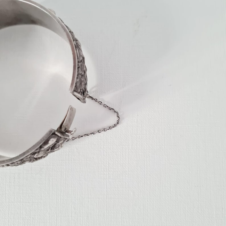 May include: A silver-toned hinged cuff bracelet featuring a textured, embossed surface pattern. The bracelet includes a secure clasp mechanism and a delicate, thin metal safety chain attached to both sides to prevent the piece from falling off.