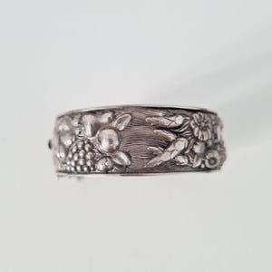 May include: A silver-toned metal cuff bracelet featuring a detailed embossed design of grapes, wheat stalks, and small flowers. The surface has a textured, oxidized finish that highlights the intricate botanical relief patterns across the band.
