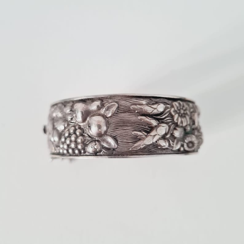 May include: A silver-toned metal cuff bracelet featuring a detailed embossed design of grapes, wheat stalks, and small flowers. The surface has a textured, oxidized finish that highlights the intricate botanical relief patterns across the band.