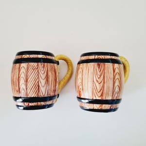 Könnte beinhalten: Zwei Keramikbecher mit einem Fassdesign. Die Becher haben ein hellbraunes Holzmaserungsmuster mit schwarzen Bändern und einem gelben Griff. Die Becher sind so gestaltet, dass sie wie Miniaturfässer aussehen.