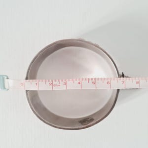 May include: A circular silver-toned metal container with a smooth interior surface. A white measuring tape is positioned across the center, indicating a diameter of approximately 8.5 centimeters for the circular opening of the container.