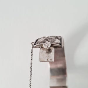 May include: A silver cuff bracelet featuring an intricate, embossed floral design on the clasp area. The bracelet includes a delicate attached safety chain and a rectangular metal tab with small, stamped hallmarks visible on the surface.