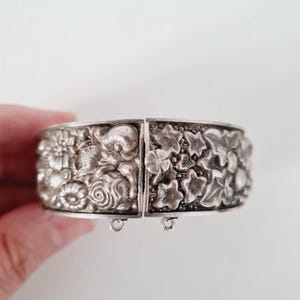 Puede incluir: Un brazalete rígido articulado de tono plateado con un diseño en relieve detallado de varias flores, capullos redondeados y hojas de hiedra texturizadas. La pulsera cuenta con un mecanismo de cierre central con un pequeño detalle floral.