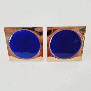 Puede incluir: Un par de gemelos dorados con forma cuadrada. Cada gemelo presenta un centro circular azul intenso, brillante y vibrante. El diseño es simple pero elegante, adecuado para vestimenta formal o para agregar un toque de color a un atuendo.