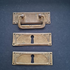 May include: Three antique brass drawer pulls and keyhole covers. The top piece is a drawer pull with a curved handle and two round knobs. Below are two keyhole covers with a decorative leaf design.