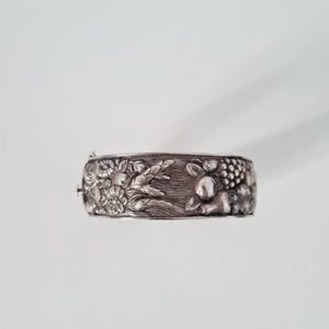 May include: A silver-toned metal hinged bangle bracelet featuring a detailed embossed relief pattern. The design displays a collection of various flowers, wheat stalks, and clusters of grapes, all rendered in a metallic finish with dark accents.