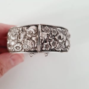 May include: A silver-toned hinged cuff bracelet featuring a detailed relief pattern of various flowers on one side and a dense arrangement of ivy leaves on the other. The piece includes a small flower-shaped clasp and two small metal loops at the base.