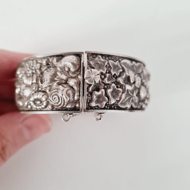 May include: A silver-toned hinged cuff bracelet featuring a detailed relief pattern of various flowers on one side and a dense arrangement of ivy leaves on the other. The piece includes a small flower-shaped clasp and two small metal loops at the base.