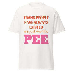 Puede incluir: Camiseta blanca con el texto "TRANS PEOPLE HAVE ALWAYS EXISTED we just want to PEE" en letras de bloque naranja y rosa. La camiseta es de estilo casual y unisex.