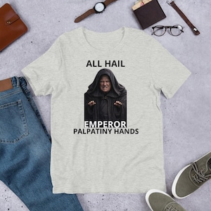 Pode incluir: Camiseta cinza claro com um gráfico do Imperador Palpatine e o texto "ALL HAIL EMPEROR PALPATINY HANDS". A imagem mostra o personagem em uma túnica preta com capuz. A camisa é feita de um material macio.