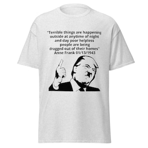 May include: Light gray t-shirt featuring a black and white graphic of a man giving a thumbs up, with a quote from Anne Frank. The quote reads: "Terrible things are happening outside at anytime of night and day poor helpless people are being dragged out of their homes" Anne Frank 01/13/1943.