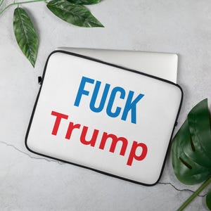 Pode incluir: Capa branca para laptop com as palavras "FUCK Trump" em letras maiúsculas azuis e vermelhas. A capa tem uma borda preta e está sobre uma superfície clara com folhas verdes.