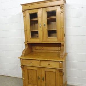 Könnte beinhalten: Ein hoher, hellfarbiger Holzschrank mit Glastüren oben und soliden Türen unten. Der Schrank hat zwei Schubladen und dekorative Schnitzereien. Das Holz hat eine natürliche Oberfläche.