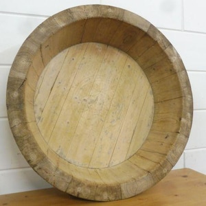 Könnte beinhalten: Ein großes, rundes Holzbecken, das aus mehreren hellen Holzplanken mit sichtbarer Maserung gefertigt ist. Das Gefäß verfügt über einen dicken, stabilen Rand und einen flachen, kreisförmigen Boden, der eine natürliche, verwitterte Textur aufweist.