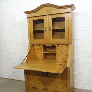 Könnte beinhalten: Ein helles Sekretär-Schreibtisch aus Holz mit einem Aufsatz. Der Schreibtisch hat eine herunterklappbare Schreibfläche, mehrere Schubladen und Schranktüren mit Glasfront. Der Schreibtisch ist aus Holz und hat eine natürliche Oberfläche.