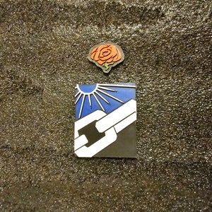 May include: Two enamel pins, one with a blue and white design of a sun and a chain link, and the other with a red rose.