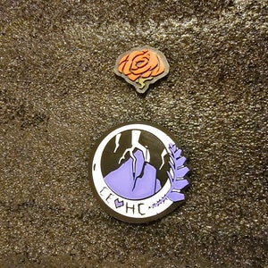 May include: Two enamel pins, one with a purple mountain and a lightning bolt, and the other with a red rose. The mountain pin has the text "LEHC" around the edge.