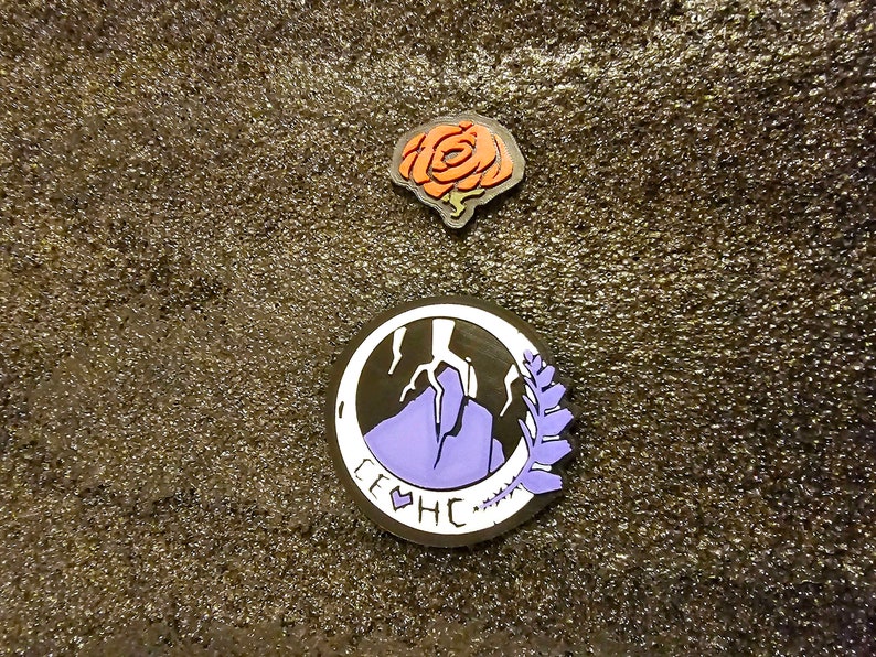 May include: Two enamel pins, one with a purple mountain and a lightning bolt, and the other with a red rose. The mountain pin has the text "LEHC" around the edge.