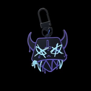 May include: A black and purple keychain with a cartoon demon face. The demon has a cross on its forehead and dripping paint on its face.