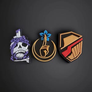 Pode incluir: Três patches decorativos com designs únicos. Um apresenta uma caveira com correntes roxas, outro uma mão segurando uma flor azul e o último um escudo com detalhes vermelhos e dourados. Todos em um fundo escuro.