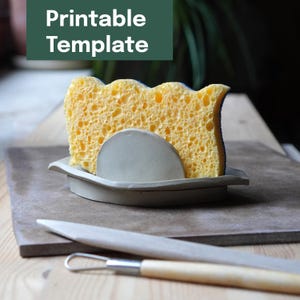 May include: A yellow sponge with a blue trim sits in a gray ceramic holder. The holder has a curved design and a half-circle support. The words "Printable Template" are in a green rectangle above the sponge.