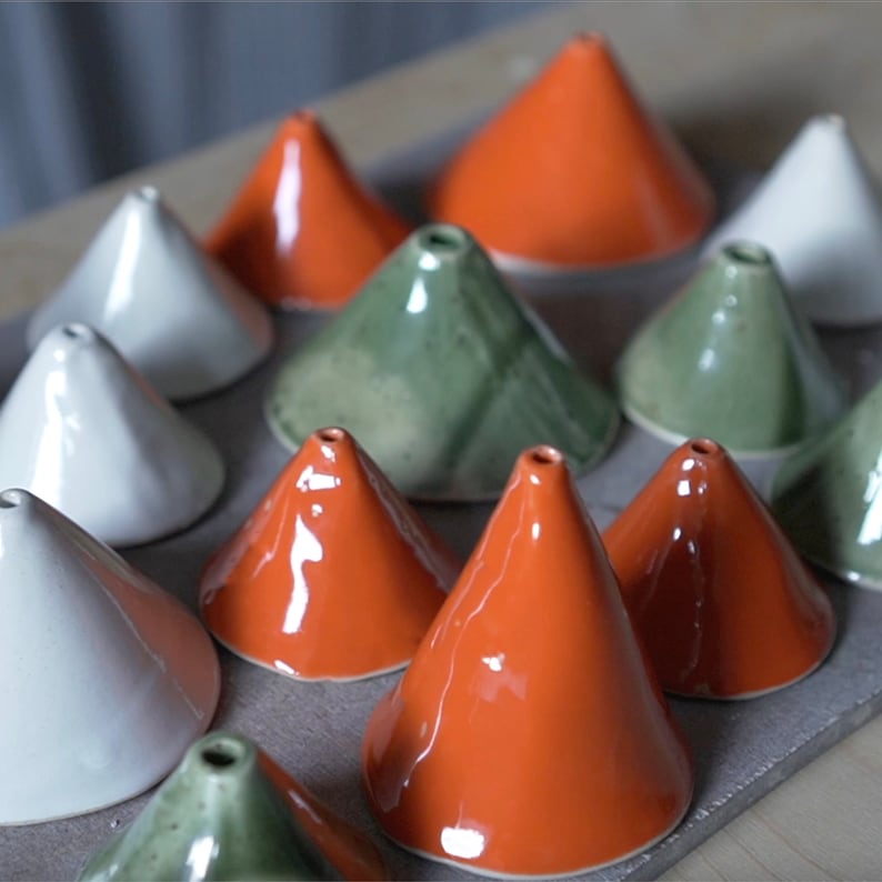 May include: A collection of ceramic cone-shaped objects in various colours, including white, green, and orange. The objects are arranged in a staggered pattern on a brown surface.
