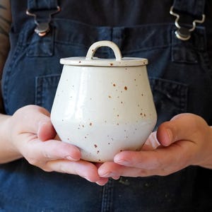 May include: A handcrafted, cream-coloured ceramic jar with a lid and a small handle. The jar features a speckled pattern with brown flecks and a glossy finish. The jar is held in both hands, highlighting its form and texture.