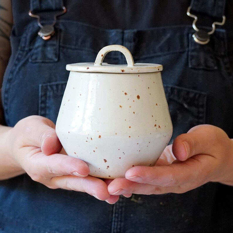 May include: A handcrafted, cream-coloured ceramic jar with a lid and a small handle. The jar features a speckled pattern with brown flecks and a glossy finish. The jar is held in both hands, highlighting its form and texture.