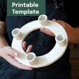 May include: A white ceramic candle holder with four cylindrical candle cups. The holder has a circular design with speckled detailing. The text "Printable Template" is in a green rectangle above the holder.