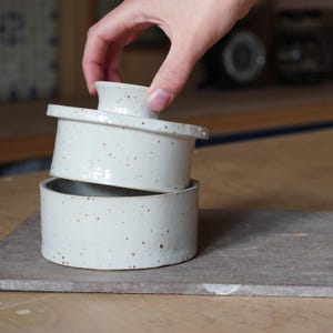 Op de afbeelding: Een witte keramische botervloot met een gespikkelde glazuur. Het deksel wordt van de basis gelicht.