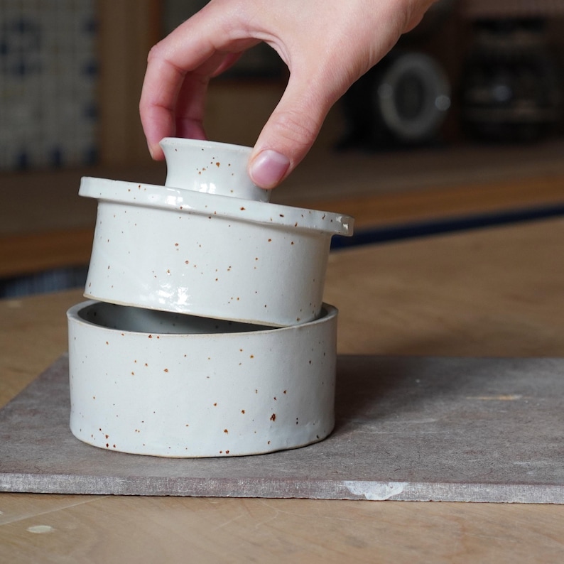 Op de afbeelding: Een witte keramische botervloot met een gespikkelde glazuur. Het deksel wordt van de basis gelicht.