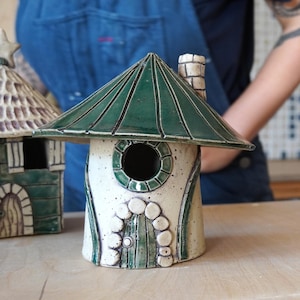 Pu&ograve; includere: Una casetta per uccelli in ceramica con un tetto verde a coste e un'apertura rotonda. La casetta &egrave; bianco sporco con dettagli verdi, tra cui un telaio di finestra circolare e una porta decorativa. &Egrave; presente un piccolo camino.