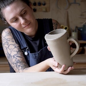 K&ouml;nnte beinhalten: Eine Person h&auml;lt eine gro&szlig;e, unfertige, hellbraune Keramiktasse mit Henkel. Die Tasse ist leicht verzogen und hat eine breite, offene Oberseite.