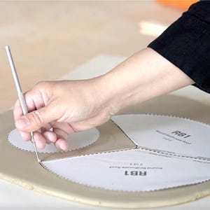 Pu&ograve; includere: Una mano usa uno strumento di metallo per tagliare intorno a un modello di carta etichettato "RB1 Round Birdhouse Roof". Il modello &egrave; su una superficie marrone chiaro. La persona indossa una camicia nera a maniche lunghe.