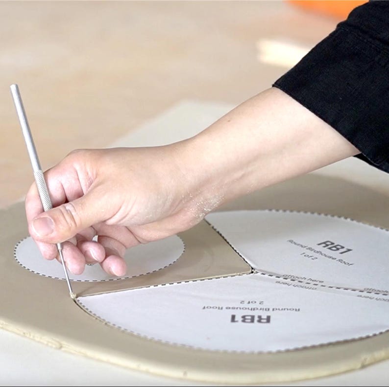 Pu&ograve; includere: Una mano usa uno strumento di metallo per tagliare intorno a un modello di carta etichettato "RB1 Round Birdhouse Roof". Il modello &egrave; su una superficie marrone chiaro. La persona indossa una camicia nera a maniche lunghe.
