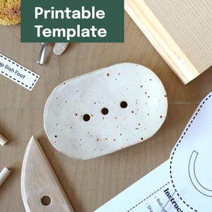 Könnte beinhalten: Eine gesprenkelte, cremefarbene Keramik-Seifenschale mit drei Abflusslöchern. Die ovale Schale ist von Bastelmaterialien und einem grünen Banner mit der Aufschrift "Printable Template" umgeben.
