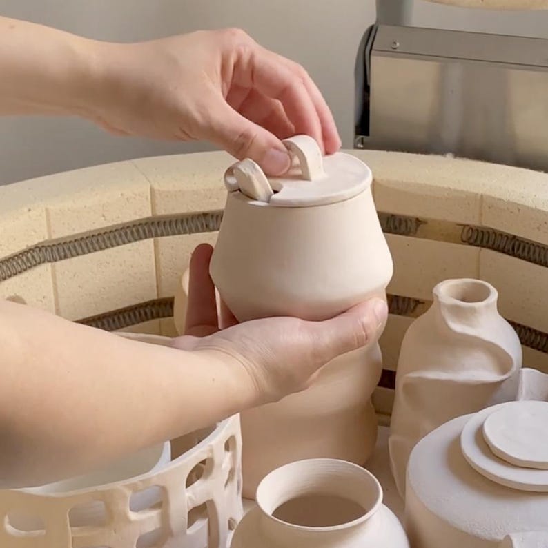 May include: A person holding a small, off-white ceramic sugar bowl with a lid and a small spoon. The bowl has a rounded shape with a handle on the lid. Other ceramic pieces are visible in the background, including a vase and a container.