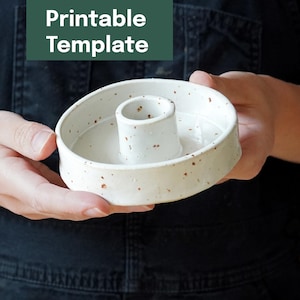 May include: A white ceramic candle holder with brown speckles and a central cylinder. The holder is round and held in a person's hands. The text "Printable Template" is in a green rectangle in the background.