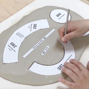 May include: Hands using a tool to cut around paper templates on a grey clay slab. The templates are labelled with text such as "Sugar Bowl Top" and "Handle". The image shows the process of creating a sugar bowl.