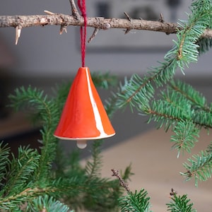 May include: A bright orange ceramic bell ornament hanging from a red string on a green pine branch.