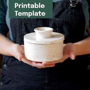 Op de afbeelding: Een witte keramische boterpot met een deksel en een klein handvat. De schaal heeft een gespikkeld ontwerp en wordt vastgehouden voor een persoon die een donkerblauwe schort draagt. De tekst "Printable Template" staat in de linkerbovenhoek.