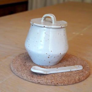 May include: A white ceramic sugar bowl with a lid and handle, speckled with brown flecks. A matching spoon rests on a round cork coaster. The sugar bowl has a unique, rounded shape. The items are set on a light-coloured wooden surface.