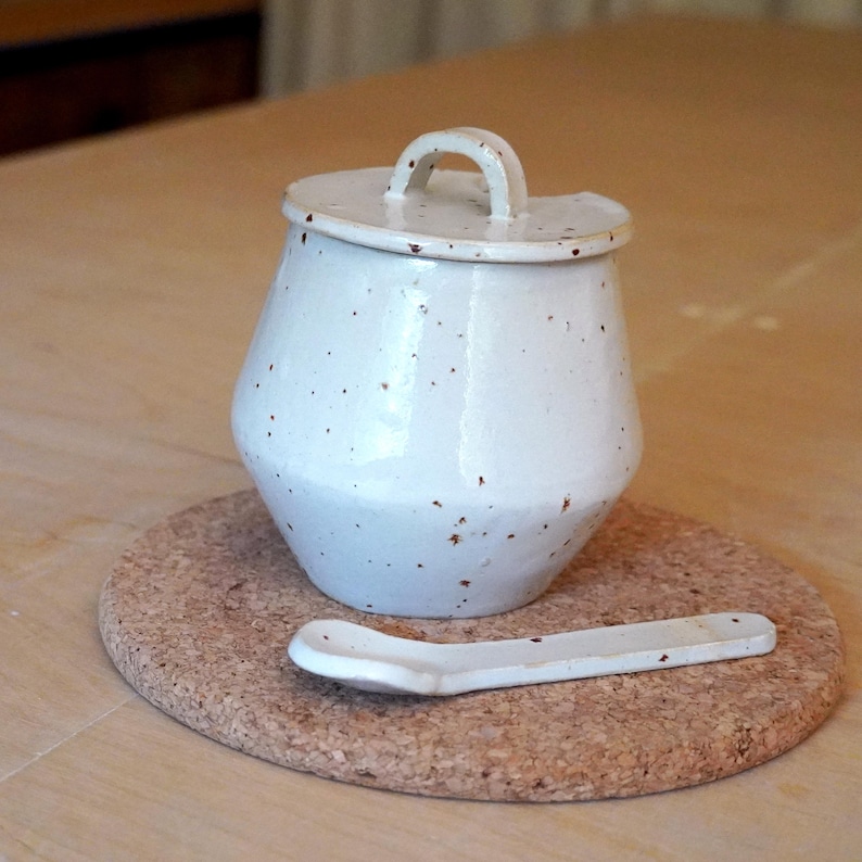 May include: A white ceramic sugar bowl with a lid and handle, speckled with brown flecks. A matching spoon rests on a round cork coaster. The sugar bowl has a unique, rounded shape. The items are set on a light-coloured wooden surface.
