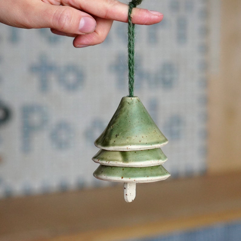 May include: A green ceramic Christmas tree bauble with a white speckled glaze. The bauble is hanging from a green string.