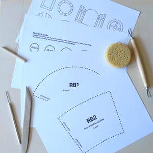 Pu&ograve; includere: Una collezione di strumenti artigianali e modelli stampati per progetti di ceramica. Include una spugna, strumenti in legno e modelli di carta con linee tratteggiate e testo, tra cui "RB1" e "RB2 Round Birdhouse Side".