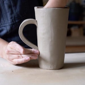 K&ouml;nnte beinhalten: Eine Hand formt eine beige Ton-Tasse mit Henkel. Die Tasse ist hoch und zylindrisch mit einem leicht ausgestellten oberen Rand.