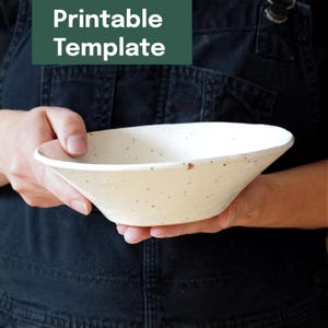 Pode incluir: Uma tigela de cerâmica creme com um design salpicado é segurada em frente a um fundo escuro. A tigela tem uma forma larga e rasa. O texto "Printable Template" está em um retângulo verde no canto superior esquerdo.