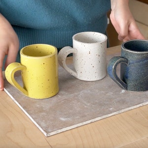 May include: Three ceramic mugs with heart-shaped handles. The mugs are speckled with white, yellow, and blue glaze.